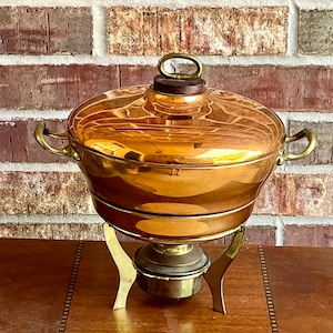 May include: A copper chafing dish with a brass stand and a wooden knob on the lid. The chafing dish is on a wooden table with a patterned inlay.