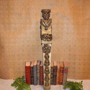 Puede incluir: Un corbel de madera pintado de oro con una cara humana tallada y detalles florales. El corbel está de pie sobre una superficie de mármol con una pila de libros delante.