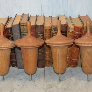 May include: Four wooden finials with a turned top and a threaded base. The finials are a light brown color and have a smooth finish. They are arranged in a row against a background of books.