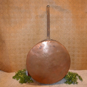 May include: A large, round copper pan with a long, metal handle. The pan is hanging from a hook on a wall.