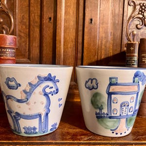 May include: Two white ceramic planters with hand-painted blue and green designs. The left planter features a blue horse with pink spots, while the right planter displays a blue house with green trees and a pink path, both with blue rim accents.