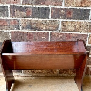 May include: A dark brown wooden magazine rack with a simple design. The rack has a rectangular shape with slightly curved sides and a flat top. It is sitting on a concrete surface.