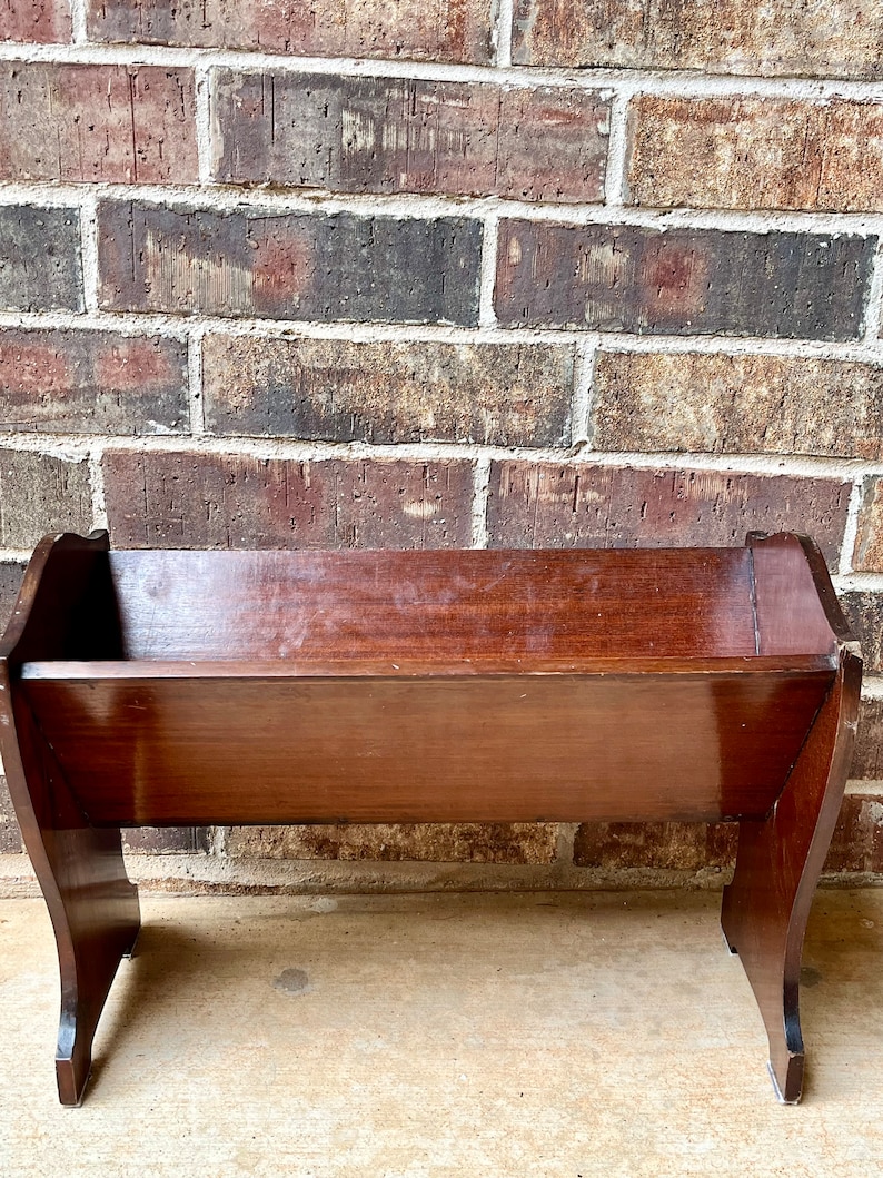 May include: A dark brown wooden magazine rack with a simple design. The rack has a rectangular shape with slightly curved sides and a flat top. It is sitting on a concrete surface.