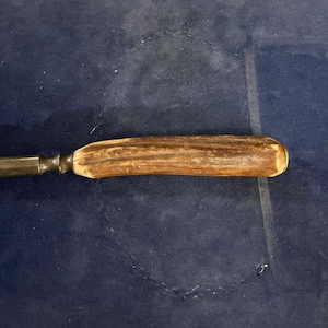 May include: A close-up of a brown antler handle attached to a metal knife blade. The knife is lying on a blue surface.