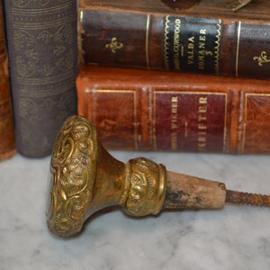 May include: A vintage brass bottle stopper with intricate floral designs and a wooden screw. The stopper is lying on a white marble surface.