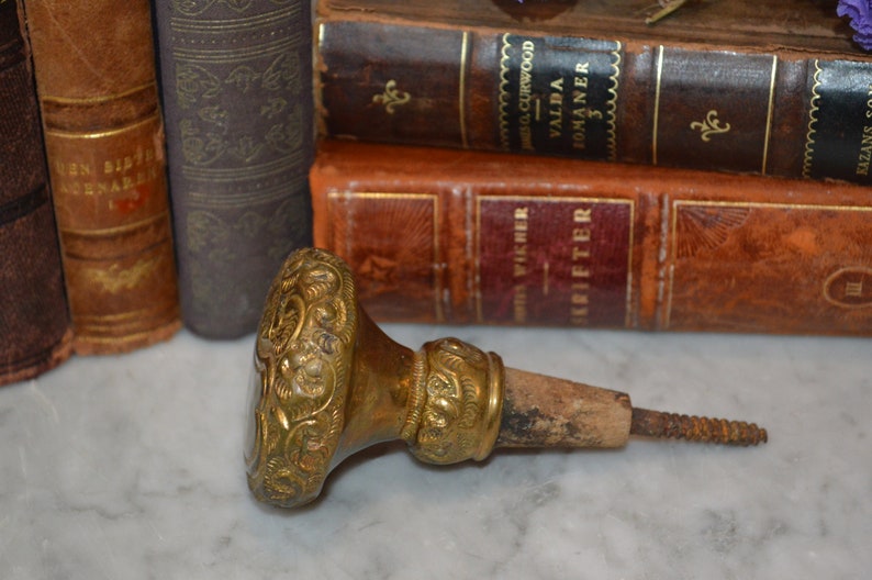 May include: A vintage brass bottle stopper with intricate floral designs and a wooden screw. The stopper is lying on a white marble surface.