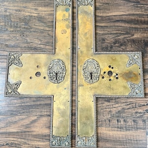 May include: Two antique brass door plates with ornate designs. The plates are rectangular and have a decorative border. The plates are separated by a small gap.