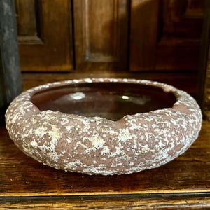 Puede incluir: Un pequeño macetero redondo de cerámica marrón con un esmalte blanco moteado. El macetero tiene una superficie rugosa y texturizada.