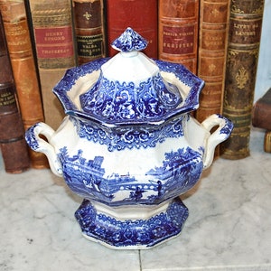 May include: A blue and white porcelain sugar bowl with a lid. The bowl has a floral design and a scene of a bridge and buildings. The bowl is sitting on a marble surface.