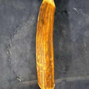 May include: A close-up of a brown antler handle for a knife or tool. The handle is smooth and polished, with a light brown color and a dark brown grain pattern.