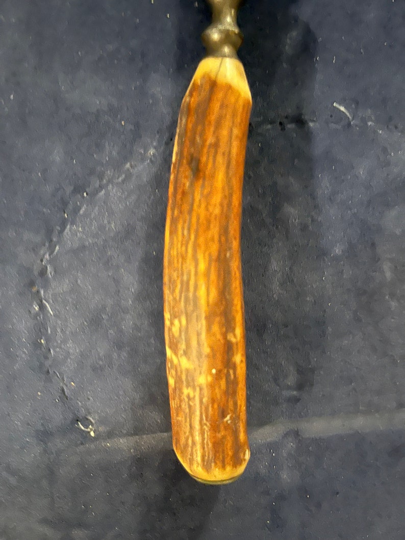 May include: A close-up of a brown antler handle for a knife or tool. The handle is smooth and polished, with a light brown color and a dark brown grain pattern.