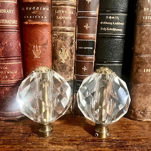 Könnte beinhalten: Zwei klare, facettierte Kristallknöpfe mit Messingbasen werden vor einer Sammlung antiker Bücher mit ledergebundenen Einbänden präsentiert. Die Bücher haben Titel in verschiedenen Sprachen und sind in Braun- und Schwarztönen gehalten.
