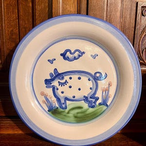 May include: A ceramic plate with a hand-painted pig design. The plate features a white center with a blue rim and a blue pig illustration with polka dots, a cloud, and flowers. The plate is approximately 10 inches in diameter.