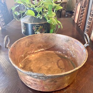 May include: A large, round copper pot with two handles. The pot has a worn, aged appearance and is sitting on a wooden table.