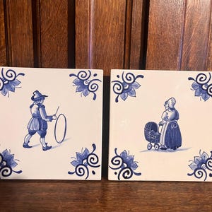 May include: Two square white ceramic tiles featuring blue hand-painted illustrations. The left tile depicts a person playing with a hoop, while the right tile shows a person standing beside a baby carriage. Both tiles have blue floral corner motifs.