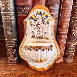 May include: A ceramic wall planter with a yellow, green, and blue floral design. A cherub with wings is painted in the center of the planter. The planter is shaped like a shell and has a small lip around the top.