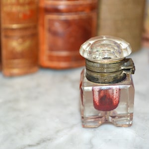 May include: A small, clear glass inkwell with a brass lid and a red inkwell inside. The inkwell is sitting on a white marble surface.