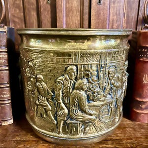 May include: A brass planter with a detailed relief depicting a historical scene with figures in period clothing. The planter has a wide, cylindrical shape and sits on a wooden surface. The background includes antique books.