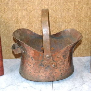 May include: An antique copper bucket with a handle and a worn patina. The bucket is sitting on a white marble surface.