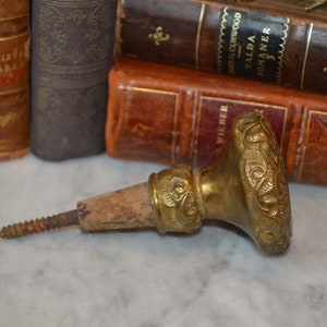 May include: A gold-toned metal bottle stopper with an ornate design. The stopper has a wooden cork and a screw-on cap.