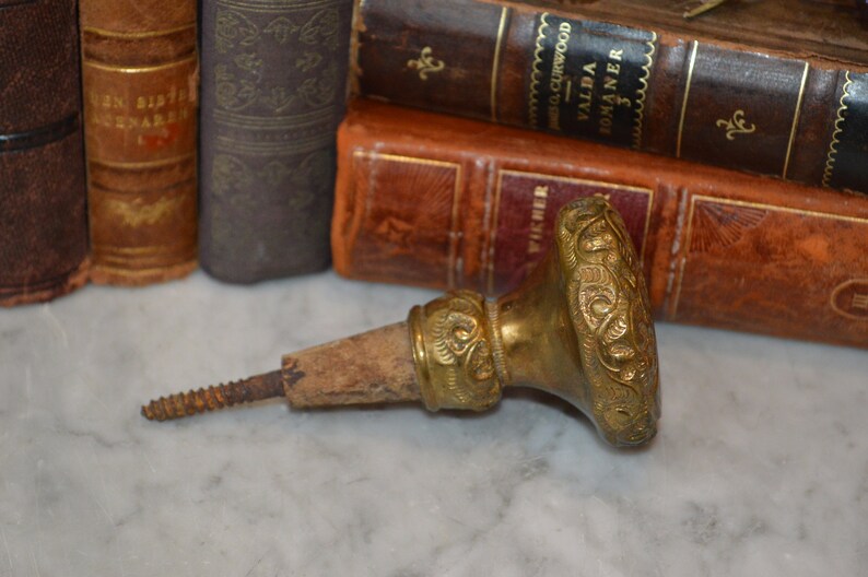 May include: A gold-toned metal bottle stopper with an ornate design. The stopper has a wooden cork and a screw-on cap.