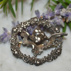 May include: Silver tone brooch featuring a cherub with wings surrounded by a wreath of flowers.