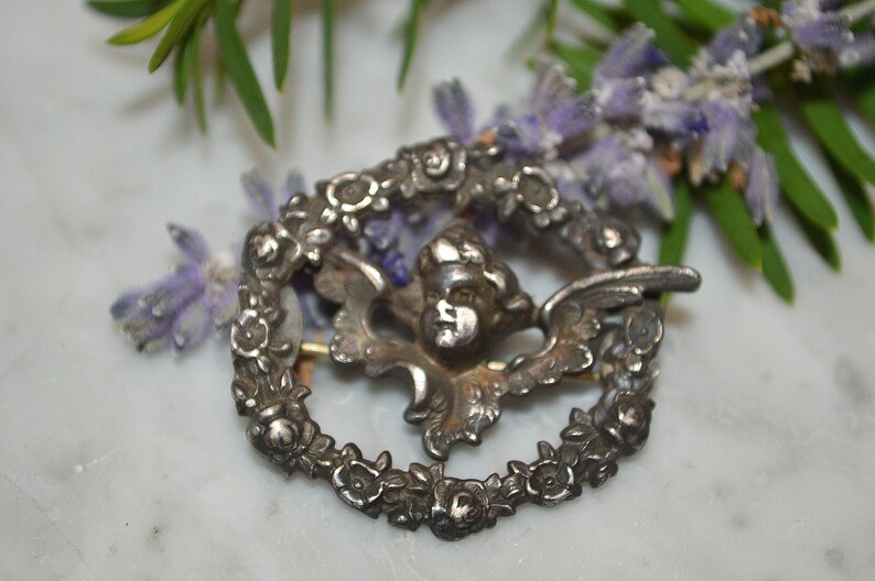 May include: Silver tone brooch featuring a cherub with wings surrounded by a wreath of flowers.