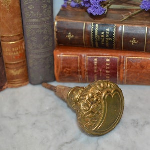 Puede incluir: Un pomo de puerta de metal dorado con intrincados diseños florales. El pomo está unido a un eje de madera. El pomo está sobre una superficie de mármol blanco.