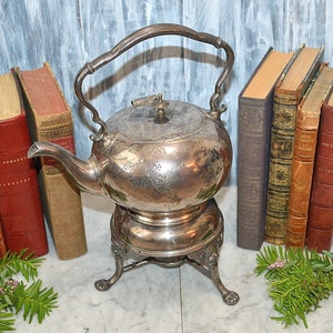 May include: Antique silver-plated tea kettle with a stand and ornate floral design. The kettle has a hinged lid and a spout. The stand has three legs and a round base.