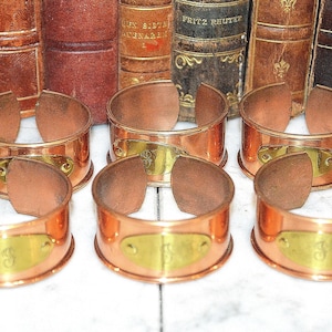 May include: Set of eight copper napkin rings with brass accents. Each ring has a rectangular brass plate with a letter engraved on it.