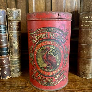 May include: A vintage red tin with a cylindrical shape, featuring a label with the text "DE ROODE PELIKAAN" and an illustration of a pelican. The tin is aged, with signs of wear and tear, and is surrounded by antique books.