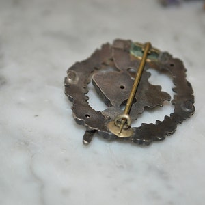 May include: Silver-toned pin with a detailed design, featuring a pin back with a safety clasp. The pin is in the shape of a wreath with a central design.