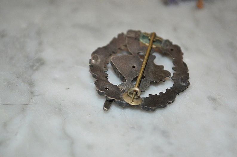May include: Silver-toned pin with a detailed design, featuring a pin back with a safety clasp. The pin is in the shape of a wreath with a central design.