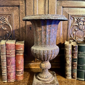 May include: A weathered, antique cast iron urn with a wide rim and pedestal base. The urn has a textured, aged appearance with rust-colored tones. Flanking the urn are several antique books with worn covers, adding to the vintage aesthetic.