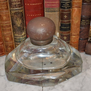 Puede incluir: Un tintero de vidrio vintage con un diseño facetado y un tapón de latón. El tintero está sobre una superficie de mármol frente a una fila de libros antiguos.