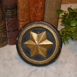 May include: A round, black wooden knob with a gold five-pointed star in the center. The knob has a gold trim around the edge.