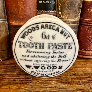 May include: A vintage white and black Woods Areca Nut Tooth Paste container. The container has a circular design with text that reads "Woods Areca Nut Tooth Paste" and "W. Woods Chemist Plymouth".