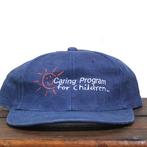 Peut inclure: Une casquette de baseball bleu marine avec un soleil orange brodé et le texte "Caring Program for Children."
