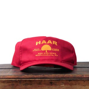 May include: A red baseball cap featuring yellow text and a tree graphic. The text reads HAAR, Wood Products, Logging Supplies, 606-549-0094, and Williamsburg, Ky. The cap has a structured front panel and a curved brim, displaying a bold colour contrast.