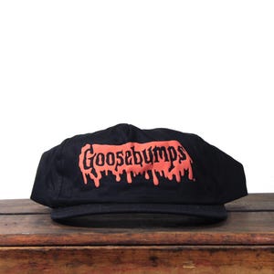 Könnte beinhalten: Schwarze Baseballkappe mit dem Wort "Goosebumps" in roter, tropfender Schrift. Die Kappe liegt auf einer Holzoberfläche. Die Kappe hat einen gebogenen Schirm und eine strukturierte Krone.