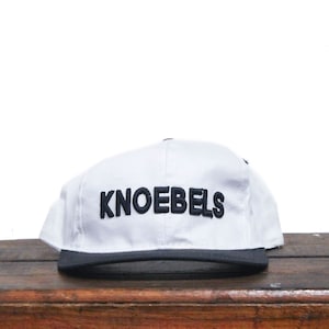 May include: White baseball cap with a black brim and black embroidered text that reads "KNOEBELS".