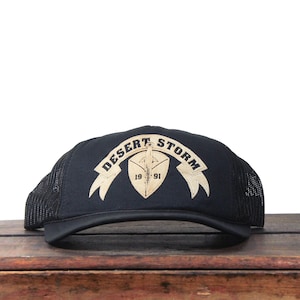Pode incluir: Boné de caminhoneiro preto com aba curva e parte traseira em rede. A frente apresenta um emblema bege com as palavras "Desert Storm" e o ano "1991". O boné está sobre uma superfície de madeira.