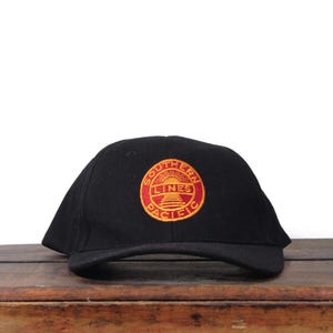 Puede incluir: Gorra de béisbol negra con un parche circular con las palabras "Southern Pacific Lines" en un diseño rojo y amarillo. La gorra está hecha de una tela oscura y se encuentra sobre una superficie de madera.