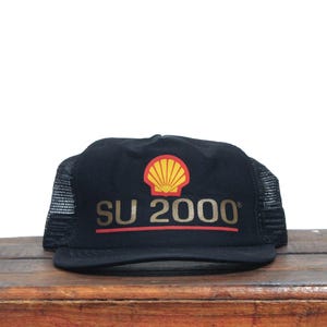 Puede incluir: Gorra de camionero negra de malla con un logotipo "SU 2000" dorado y un logotipo de Shell rojo y amarillo.