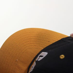 May include: A black baseball cap with a gold brim. The brim is curved and has a stitched pattern. A small gold button is on the side of the cap. A white logo is visible on the side of the cap.