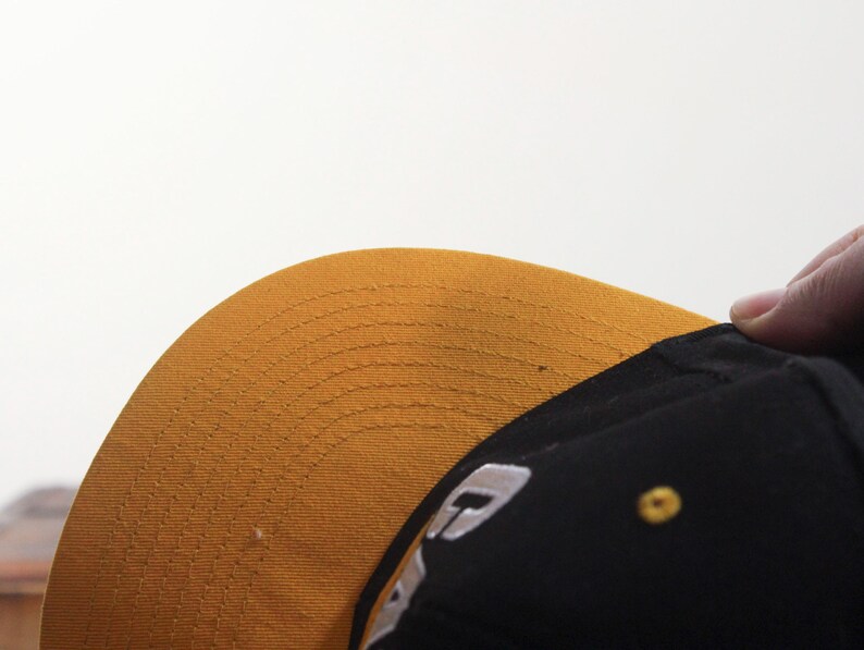 May include: A black baseball cap with a gold brim. The brim is curved and has a stitched pattern. A small gold button is on the side of the cap. A white logo is visible on the side of the cap.