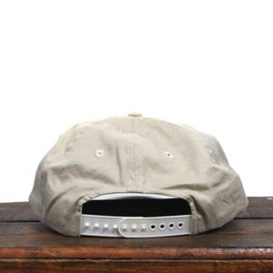 May include: A beige baseball cap with a white adjustable strap in the back. The cap is made of a soft, textured fabric.