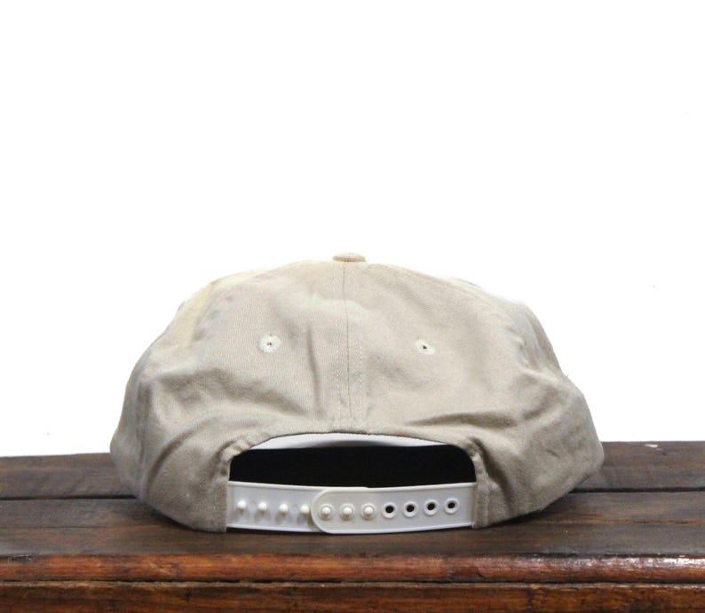 May include: A beige baseball cap with a white adjustable strap in the back. The cap is made of a soft, textured fabric.