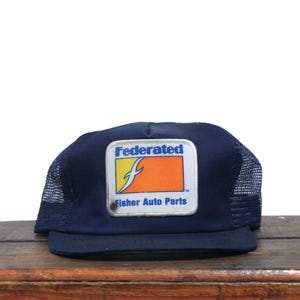 Peut inclure: Un chapeau de camionneur en maille bleu et noir avec un patch blanc, orange et jaune qui indique "Federated Fisher Auto Parts".