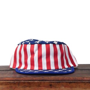 Könnte beinhalten: Eine patriotische Baseballkappe mit dem Design der amerikanischen Flagge. Die Kappe hat einen blauen Schirm mit weißen Sternen, eine blaue Oberseite mit weißen Sternen und rot-weiße Streifen. Die Kappe besteht aus Stoff und liegt auf einer Holzoberfläche.